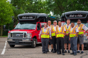 Two Metro Bus Dial-a-Ride buses parked behind a group of Metro Bus Dial-a-Ride Operators smiling.