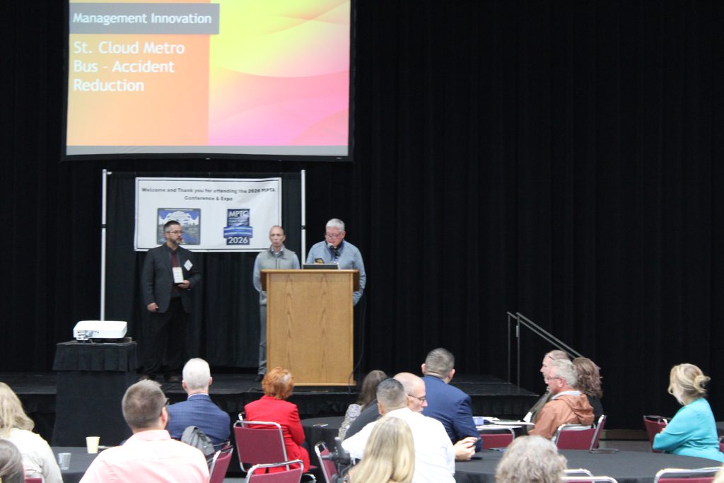 Three men on the stage behind a podium. One of them is speaking into the microphone.